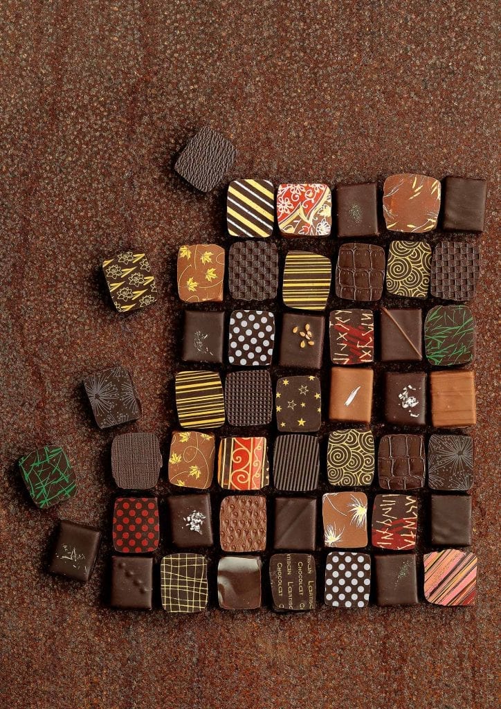 Small, decorated patterned chocolates arranged in a grid on a chocolate coloured surface similar to a mosaic.